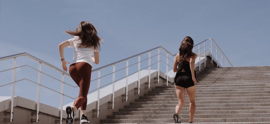 Two women running up outdoor stairs for fitness under a clear blue sky.